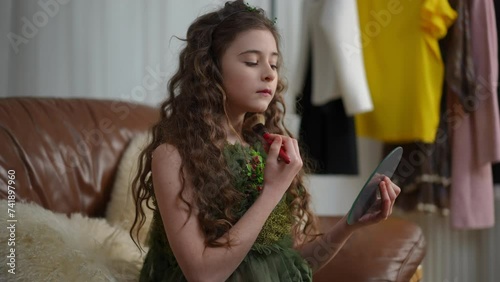 A beautiful little lady in a green dress is doing makeup, looking at herself in a small mirror that she holds in her hand. Girl preteen sitting on a sofa in a modern living room photo