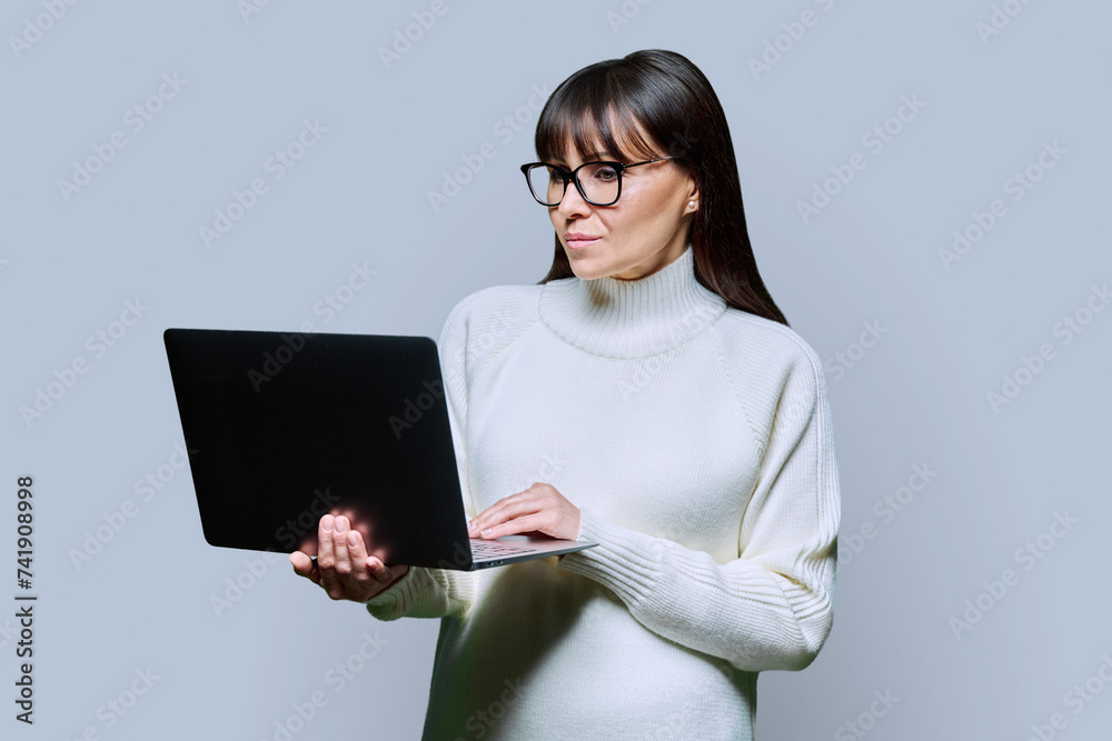 Middle-aged beautiful serious woman holding laptop on grey background Middle-aged beautiful serious woman holding laptop on grey background