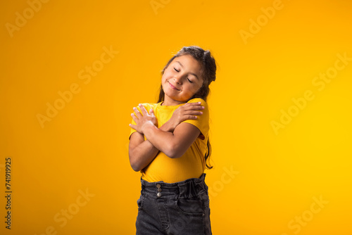 Child girl standing over isolated yellow background hugging oneself happy and positive, smiling confident. Self love and self care photo