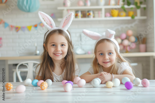 In a festive kitchen, two young girls with bunny ears sit at a table decorated with painted Easter eggs