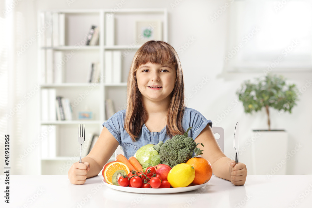 Happy little girl sitting with a plate of fruits and vegetables and holding a fork and knife