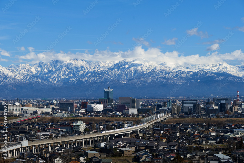 呉羽山から望む北アルプス立山連峰と富山市市街地駅を出る下り新幹線