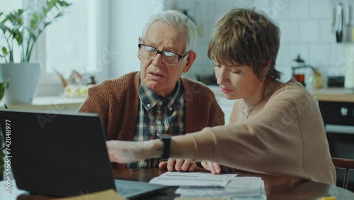 Elderly man learning how to pay bills online on laptop with assistance of young female social worker visiting him at home photo