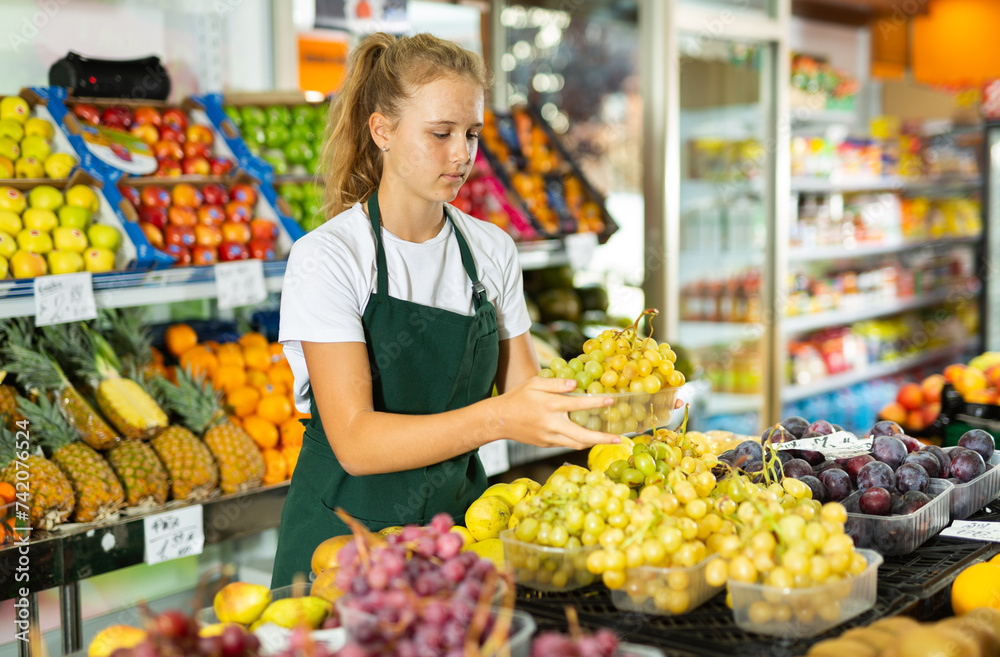 Hardworking fifteen-year-old girl who works part-time in a store as a trainee saleswoman puts grapes on the counter, laid ..out in boxes for sale