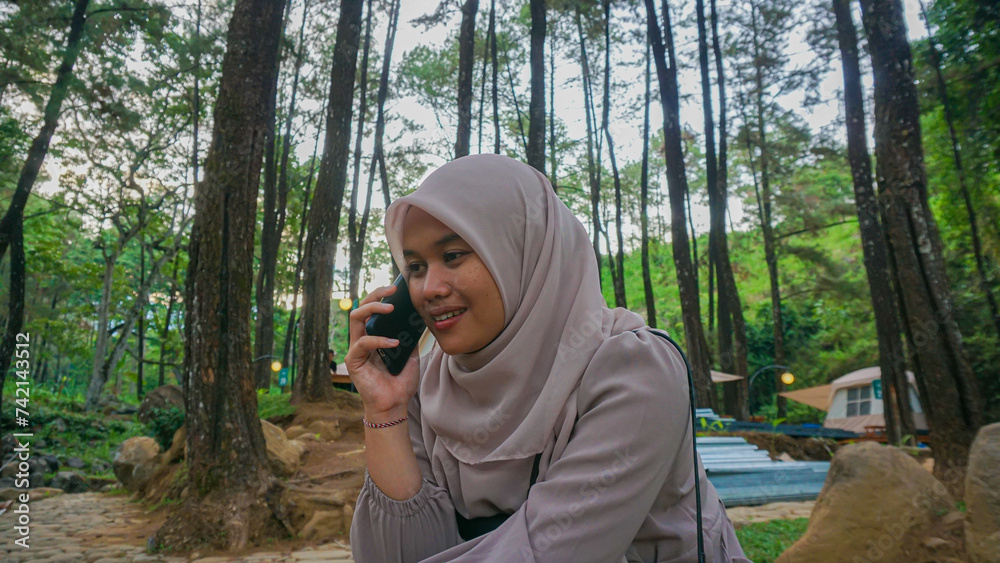 Beautiful young woman from Asia is talking on the phone while sitting under a tree in a park Beautiful young woman from Asia is talking on the phone while sitting under a tree in a park