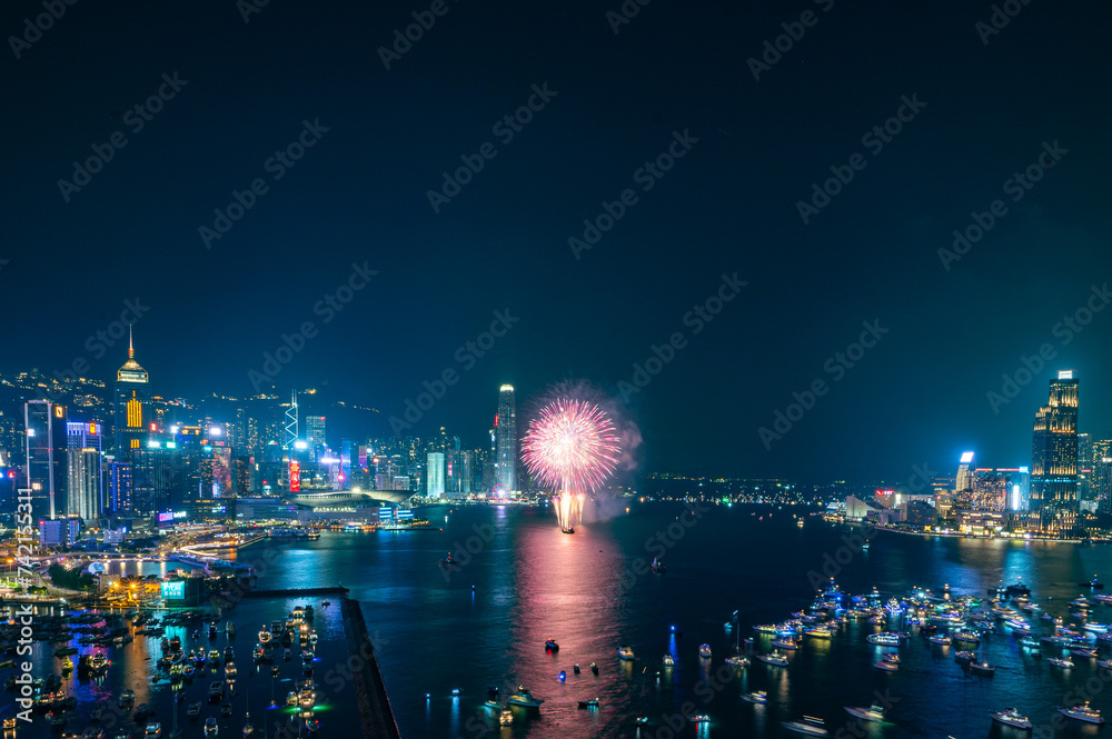 Fireworks burst brightly against the night sky above a city skyline, reflecting in the calm water below