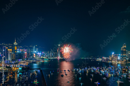 Fireworks burst brightly against the night sky above a city skyline, reflecting in the calm water below © TANG