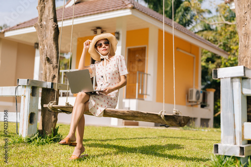 Relaxing day of an Asian female tourist wearing a hat, glasses, and dress, sitting on a swing on the lawn in front of the room Have a laptop to use for business during the summer holidays photo