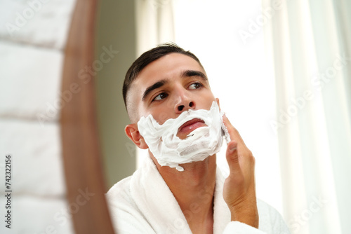 Reflection of a young man shaving in bathroom mirror close up