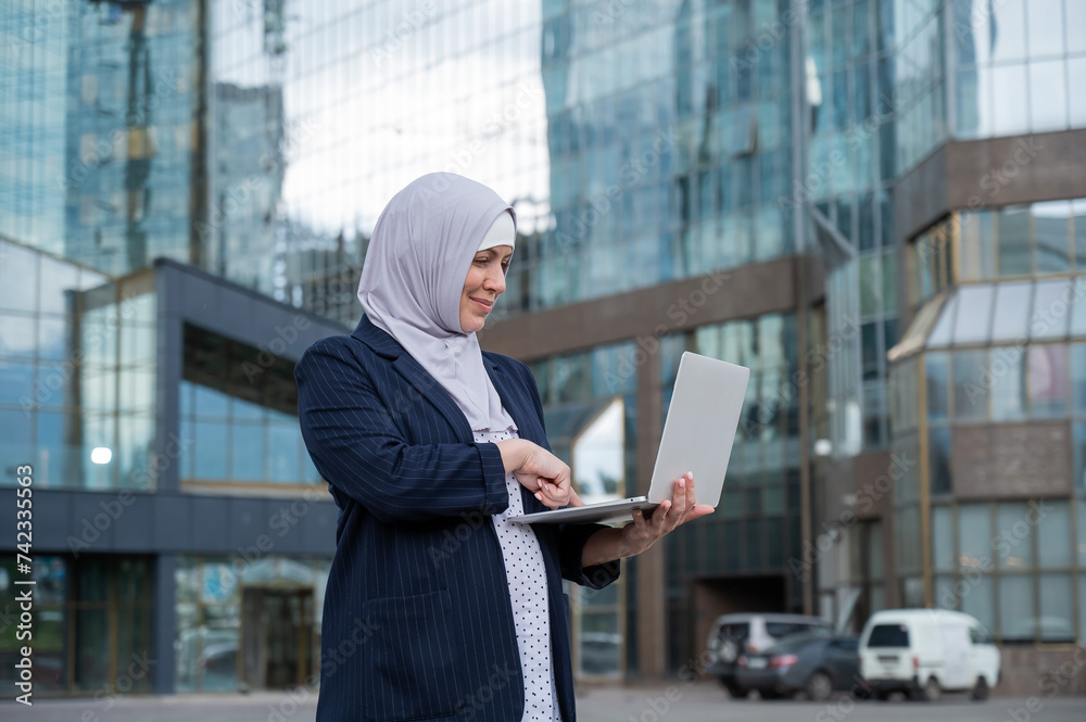 Fototapeta premium Thoughtful business woman in hijab and suit is holding a laptop outdoors.