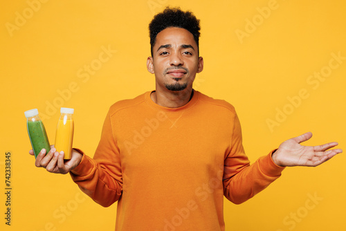 Young sad man wear casual clothes hold fruit juice green vegetable smoothie as detox diet spread hand isolated on plain yellow background. Proper nutrition healthy fast food unhealthy choice concept.