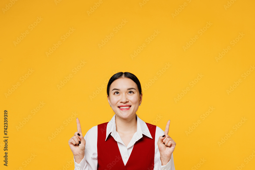 Young fun corporate lawyer employee business woman of Asian ethnicity wear formal red vest shirt work at office point index finger overhead on area isolated on plain yellow background. Career concept. Young fun corporate lawyer employee business woman of Asian ethnicity wear formal red vest shirt work at office point index finger overhead on area isolated on plain yellow background. Career concept.