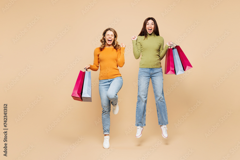 Full body young friends two women wear orange shirt casual clothes hold shopping paper package bags jump high do winner gesture isolated on plain beige background. Black Friday sale buy day concept. Full body young friends two women wear orange shirt casual clothes hold shopping paper package bags jump high do winner gesture isolated on plain beige background. Black Friday sale buy day concept.
