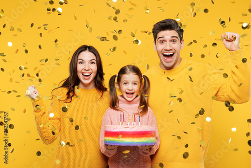 Young overjoyed happy parents mom dad with child kid girl 7-8 years old wear pink sweater casual clothes hold birthday cake do winner gesture isolated on plain yellow background. Family day concept. photo