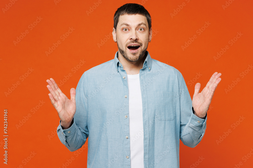 Young surprised shocked fun cool stunned man he wearing blue shirt white t-shirt casual clothes look camera spreading hands isolated on plain red orange background studio portrait. Lifestyle concept.