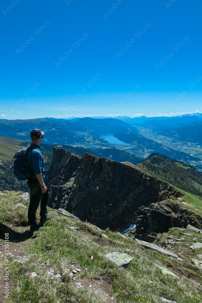 Hiker man on idyllic hiking trail on alpine meadow with scenic view of lake Millstatt seen from mountain peak Boese Nase, Ankogel Group, Carinthia, Austria. Remote landscape. Austrian Alps in summer
