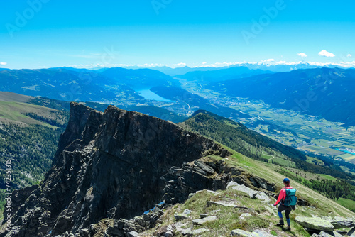 Hiker woman on idyllic hiking trail on alpine meadow with scenic view of lake Millstatt seen from mountain peak Boese Nase, Ankogel Group, Carinthia, Austria. Remote landscape. Austrian Alps in summer