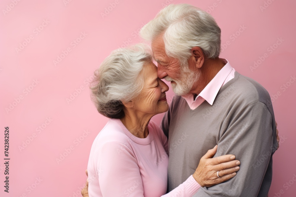
Affectionate elderly pair, both in their late 70s, European, sharing a tender embrace with eyes closed against a soft pink pastel background with copy space