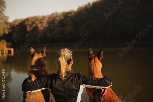 Canvas Print woman hugging two belgian shepherd malinois dogs next to a forest lake at sunset