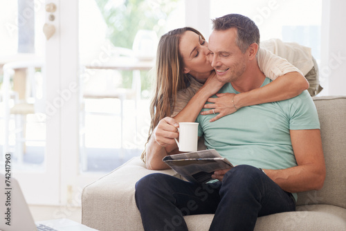 Happy couple, kiss and newspaper with coffee in home, romantic and bonding together with love in marriage. Man, woman and surprise by touch with relax, warm drink and care by sofa in modern apartment