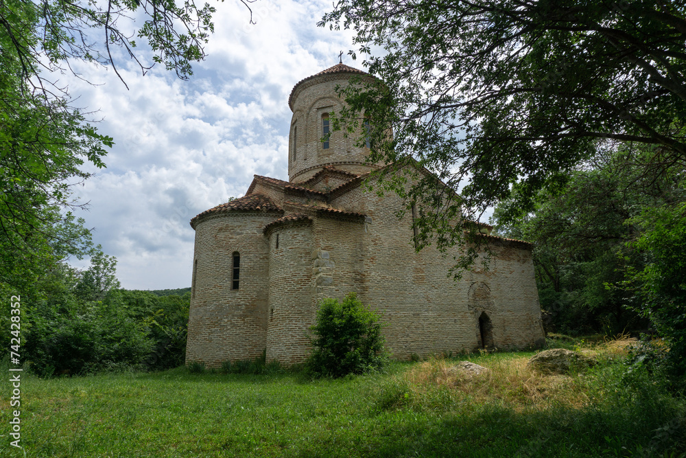 Ancient church in the forest. Bricks and stone wall. Partially destroyed arched extension at the entrance. Orange tiled roof, grass lawn. Ancient church in the forest. Bricks and stone wall. Partially destroyed arched extension at the entrance. Orange tiled roof, grass lawn.
