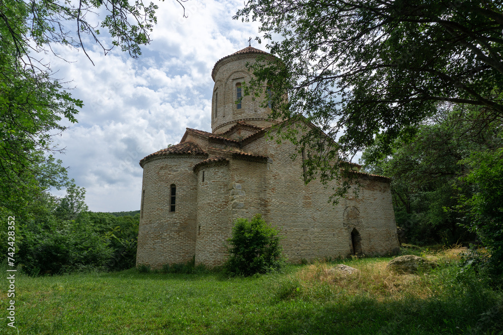 Ancient church in the forest. Bricks and stone wall. Partially destroyed arched extension at the entrance. Orange tiled roof, grass lawn.