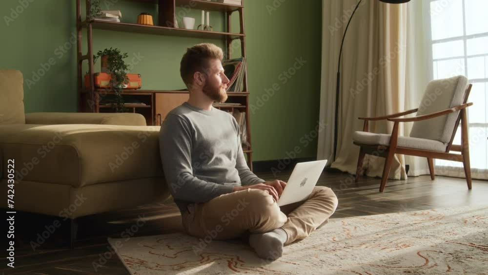 bearded man sits on floor near couch at home in living room at sunlight, working. freelancer used laptop, typing on keyboard, stops for think and look through window. bearded man sits on floor near couch at home in living room at sunlight, working. freelancer used laptop, typing on keyboard, stops for think and look through window.
