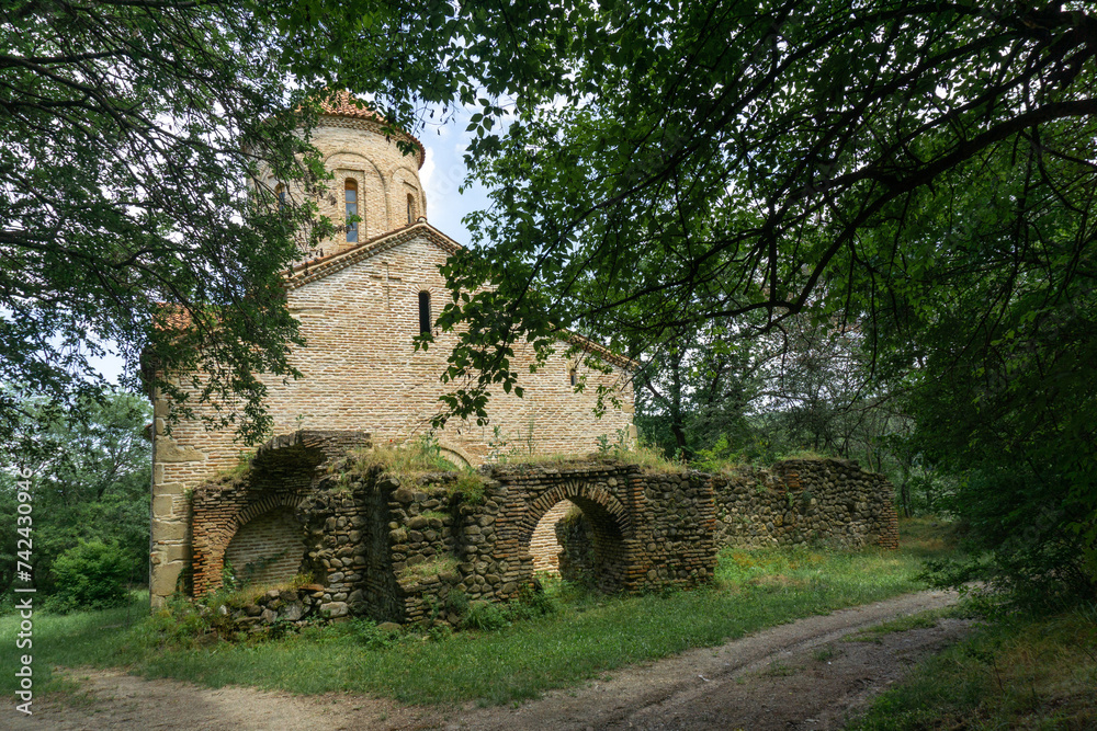 Ancient church in the forest. Bricks and stone wall. Partially destroyed arched extension at the entrance. Orange tiled roof. Grass lawn, traces of an earthen road.