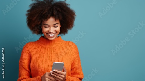 A cheerful smiling African American woman wearing an orange sweater and using a smartphone on a blue background with copy space. Positive Emotions, Facial expressions, Modern technology concepts