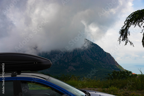 A blue car with a black roof rack stands against the backdrop of a mountain lake with mountains and clouds.