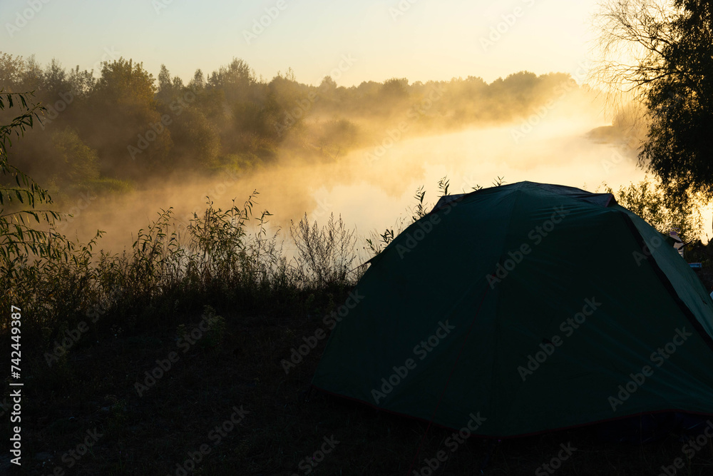 Obraz premium Green tourist tent by the river at sunrise, with morning autumn fog on the water. Outdoor tourist landscape.