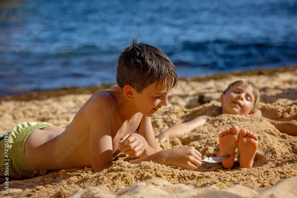 Child, tickling sibling on the beach on the feet with feather, kid cover in sand, smiling, laughing Child, tickling sibling on the beach on the feet with feather, kid cover in sand, smiling, laughing