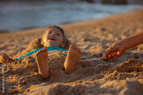 Child, tickling sibling on the beach on the feet with feather, kid cover in sand, smiling, laughing photo
