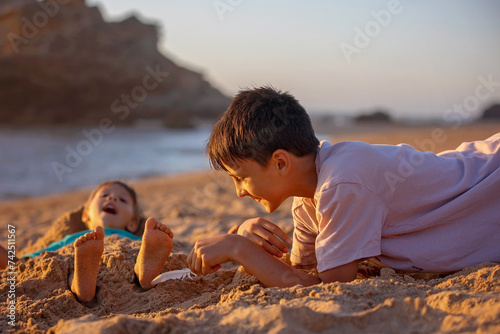 Child, tickling sibling on the beach on the feet with feather, kid cover in sand, smiling, laughing photo
