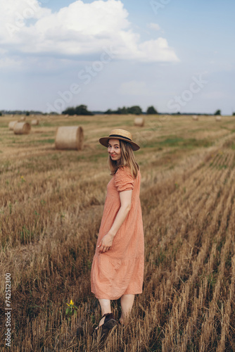 A beautiful girl in a long dress and long hair walks through a field of mown wheat. Against the background of blue sky and clouds