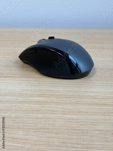 A black and gray computer mouse sits on a smooth wooden desk. The mouse is ergonomically designed and has a scroll wheel and two buttons. The desk is a light brown color.