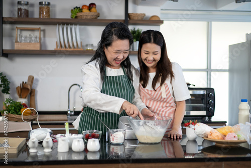 Asian female baker pastry bakery chef daughter wears apron standing smiling hugging old senior mother when mixing blending flour powder in glass bowl preparing homemade cake dough in home kitchen 
