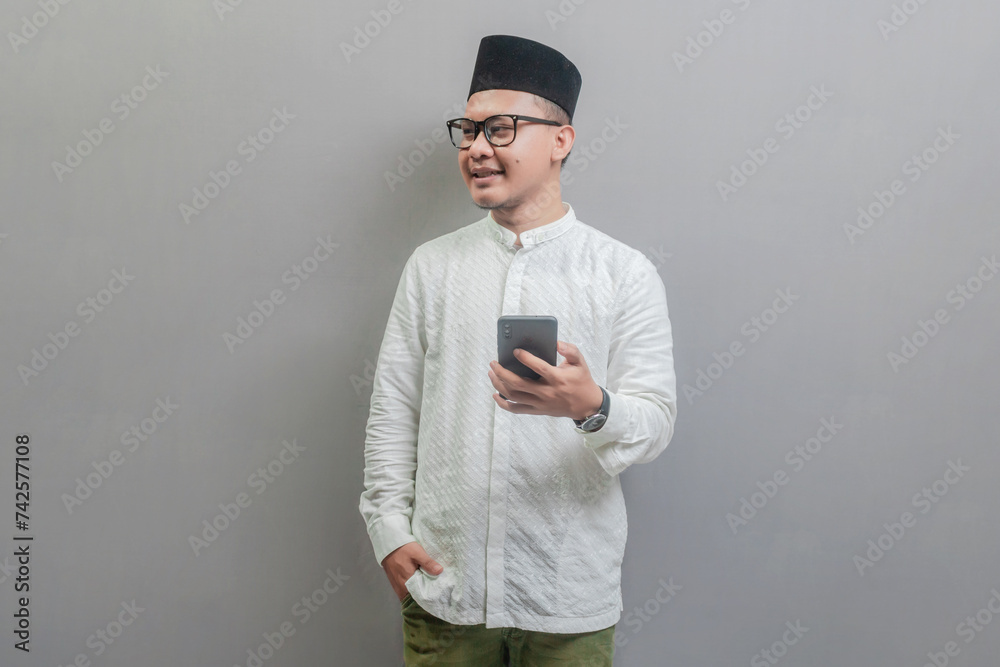 Portrait of an Asian Muslim man wearing a koko shirt and peci with shades of the fasting month, smilling and looking free space and hold his smartphone, isolated on a white background