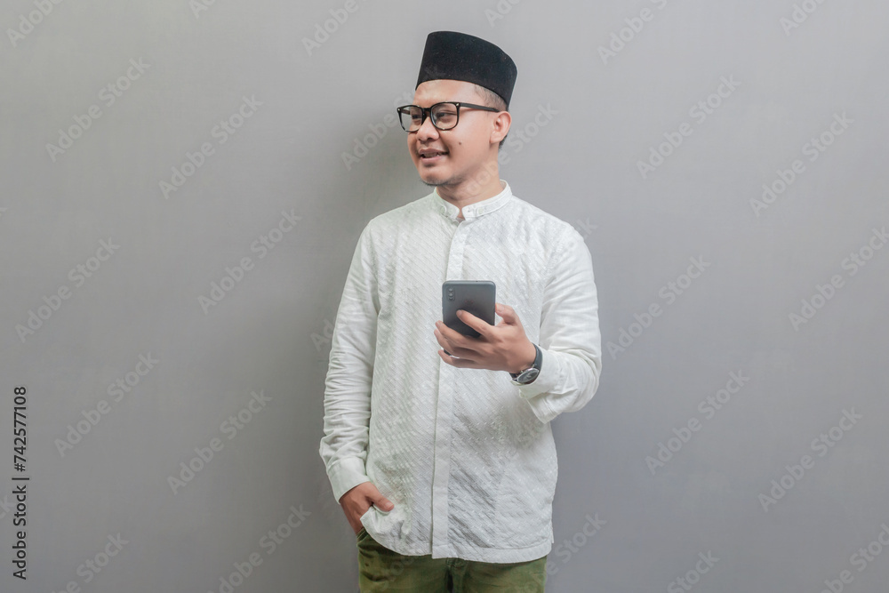 Portrait of an Asian Muslim man wearing a koko shirt and peci with shades of the fasting month, smilling and looking free space and hold his smartphone, isolated on a white background Portrait of an Asian Muslim man wearing a koko shirt and peci with shades of the fasting month, smilling and looking free space and hold his smartphone, isolated on a white background