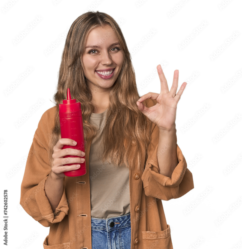 Blonde woman holding ketchup bottle in studio setting cheerful and confident showing ok gesture. Blonde woman holding ketchup bottle in studio setting cheerful and confident showing ok gesture.