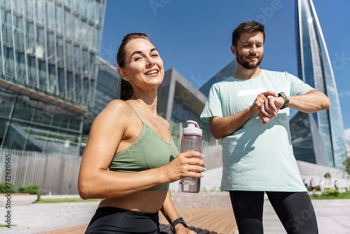 Fit couple taking a break during an outdoor workout, staying hydrated and checking time.