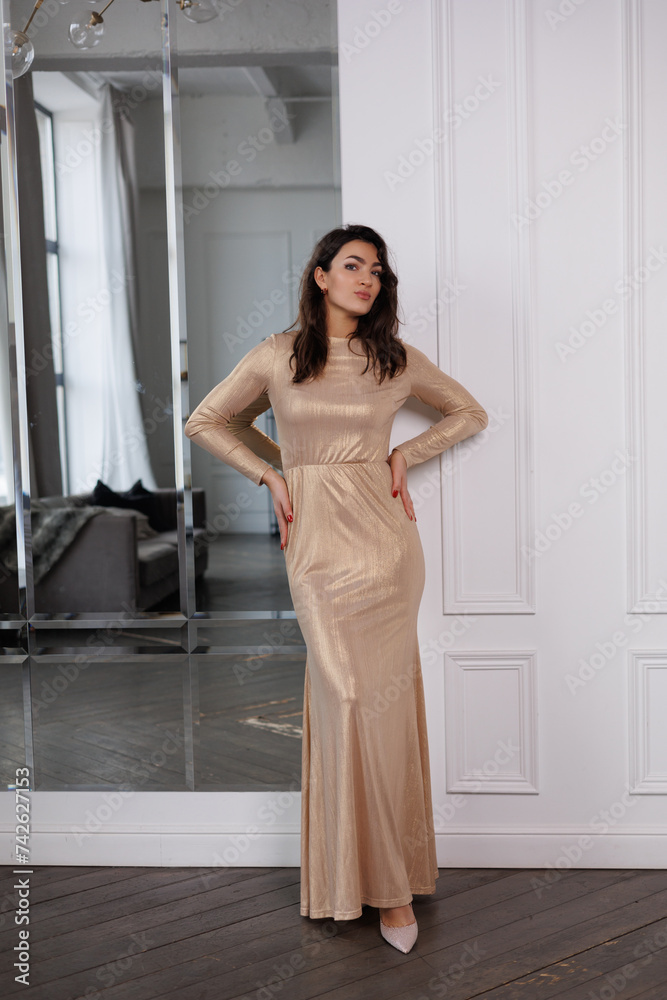 Beautiful brunette woman in long golden dress stand near mirror in room. Portrait of pretty elegant tall woman with long black hair, smile. Beautiful brunette woman in long golden dress stand near mirror in room. Portrait of pretty elegant tall woman with long black hair, smile.