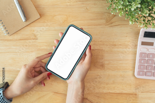 Female hands holding smartphone with mockup of blank screen on desk in office room.