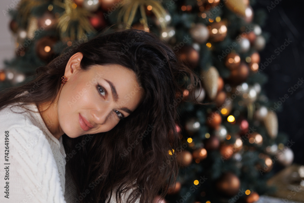 Portrait of pretty brunette woman near New Year Tree. Christmas holyday. Happy girl in cosy sweater sitting on beige sofa, smile. Celebrate New Year Eve. Woman at home relax Portrait of pretty brunette woman near New Year Tree. Christmas holyday. Happy girl in cosy sweater sitting on beige sofa, smile. Celebrate New Year Eve. Woman at home relax