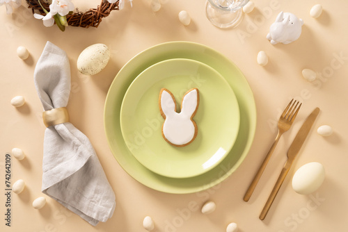 Easter table setting with white and green plate, bunny cookies, eggs on beige background. View from above. Happy Easter.