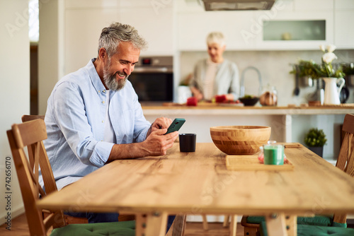 A joyful senior adult man sitting at a dining table and using his phone while waiting for the breakfast that his wife was preparing in the kitchen
