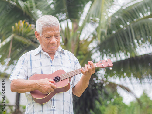 An elderly Asian man playing the ukulele, smiling and looking at the ukulele while standing in a garden.