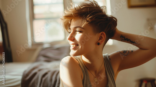 Smiling queer woman sitting, touching hair, tattoo visible, sunlit room.