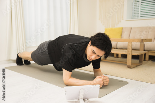 ソファのある部屋で床にヨガマットを敷いてタブレットを見て筋トレのプランクをする20代〜30代の男性