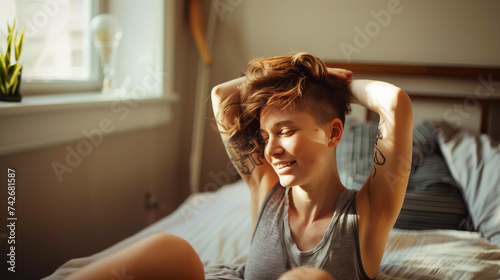 Smiling queer person sitting, stretching, touching hair, tattoo visible, sunlit room. photo